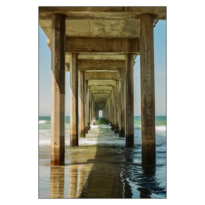 The concrete pillars of a pier extend into the distance over the ocean with waves crashing on the shore.