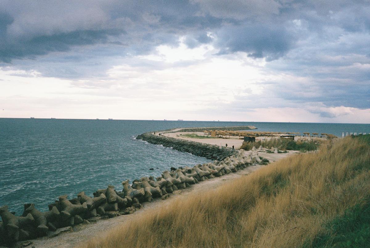 A long concrete pier with a beach and dry grass on the right extends into the sea under a cloudy sky.