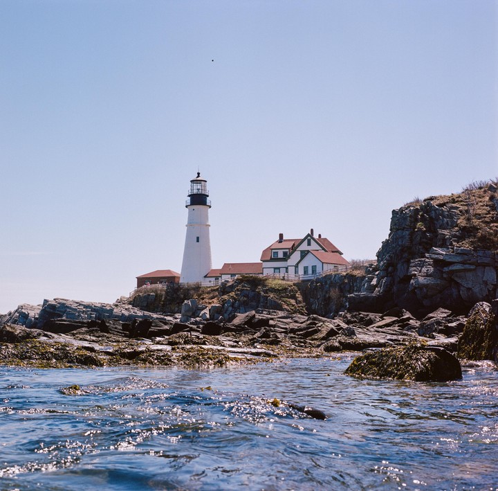A white lighthouse and buildings with red roofs stand on a rocky shore next to the ocean.