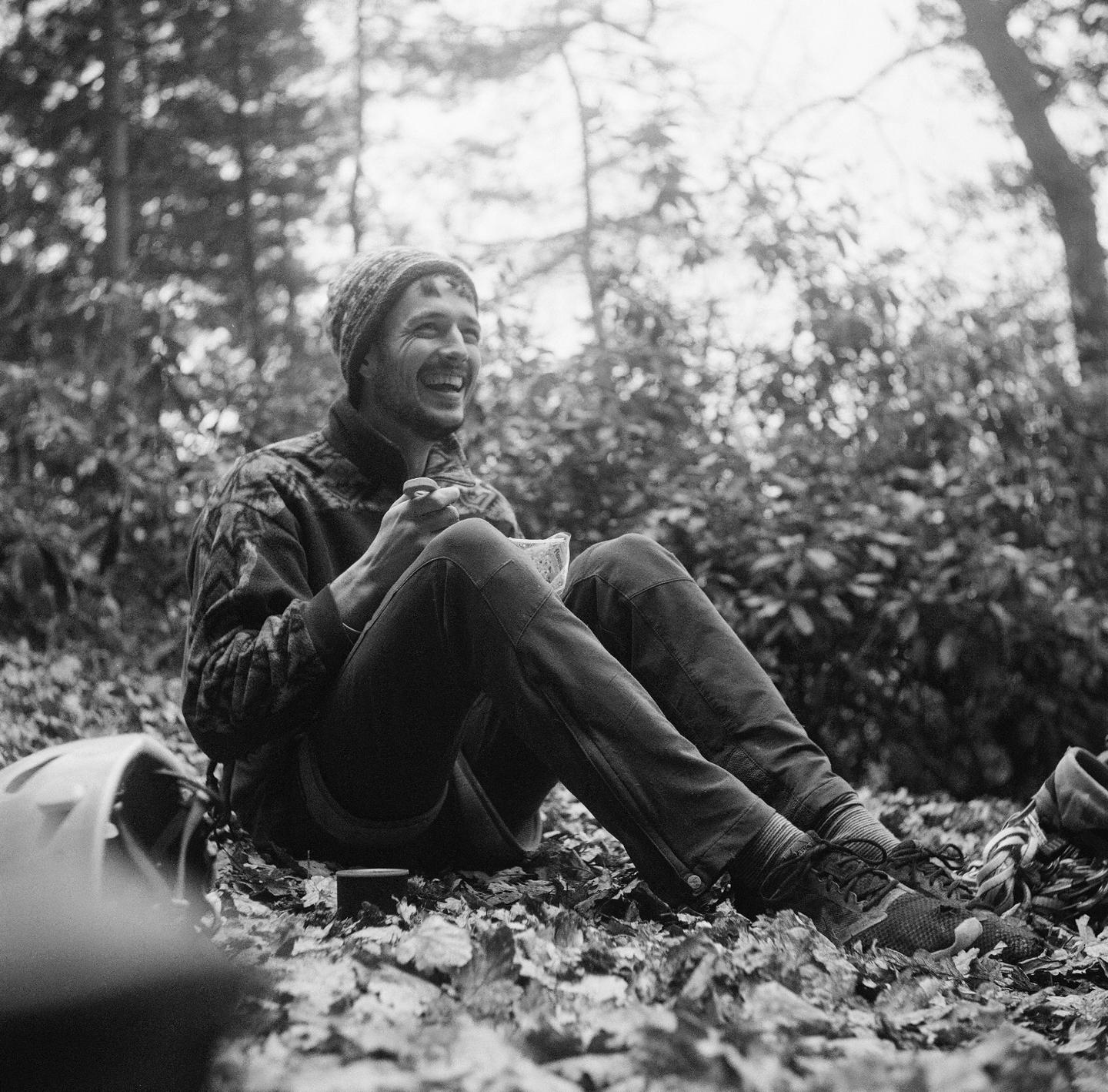 A man sits on a forest floor covered in autumn leaves, laughing while holding a small object in his hands.