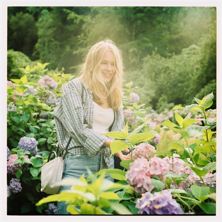 A young woman with long blonde hair smiles while standing among purple and pink hydrangea blossoms.