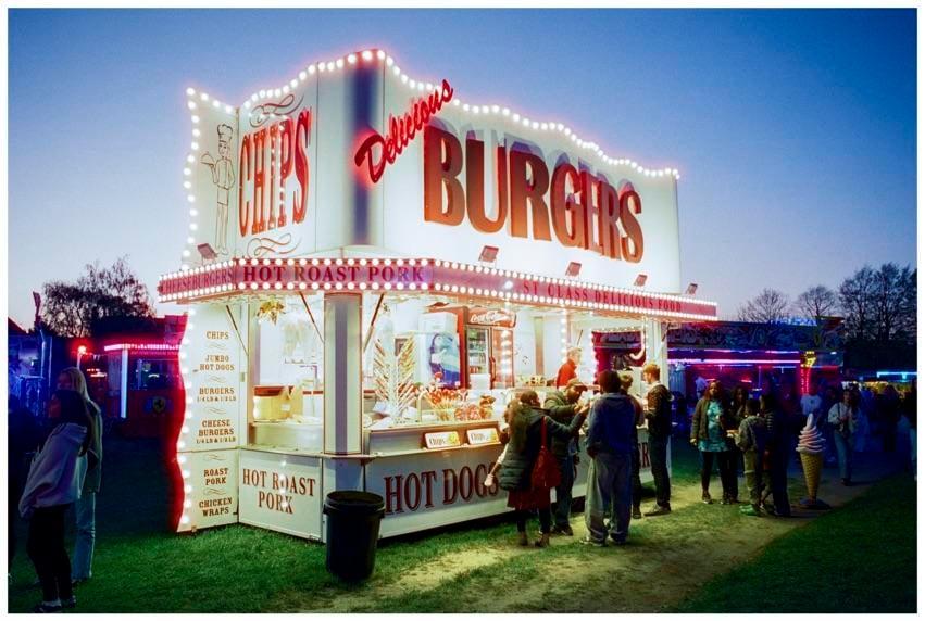 A food stall with bright lights and signs for burgers, chips, and hot dogs is surrounded by people at dusk.