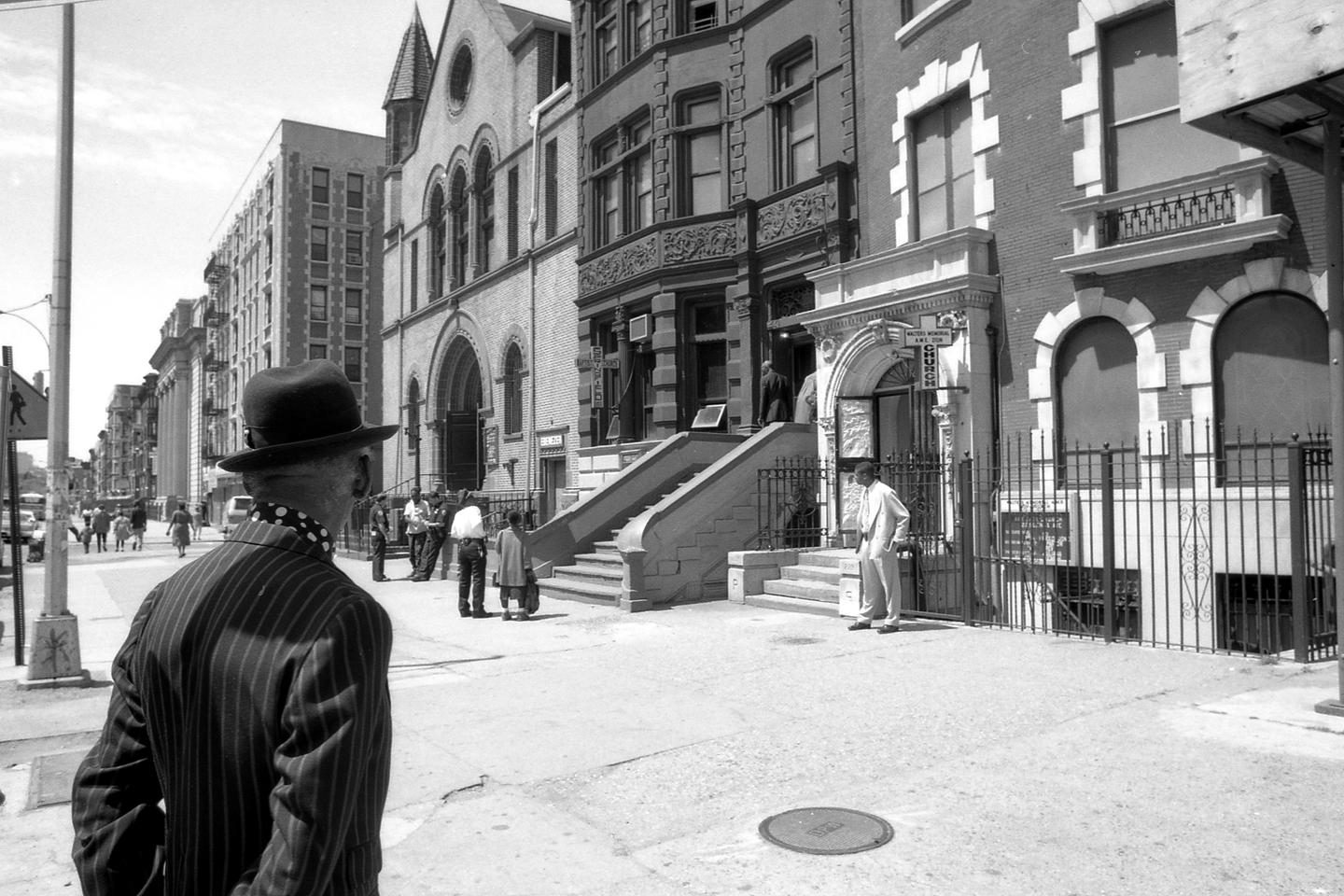 A man in a pinstripe suit and hat stands on a city street looking towards a church and other buildings.