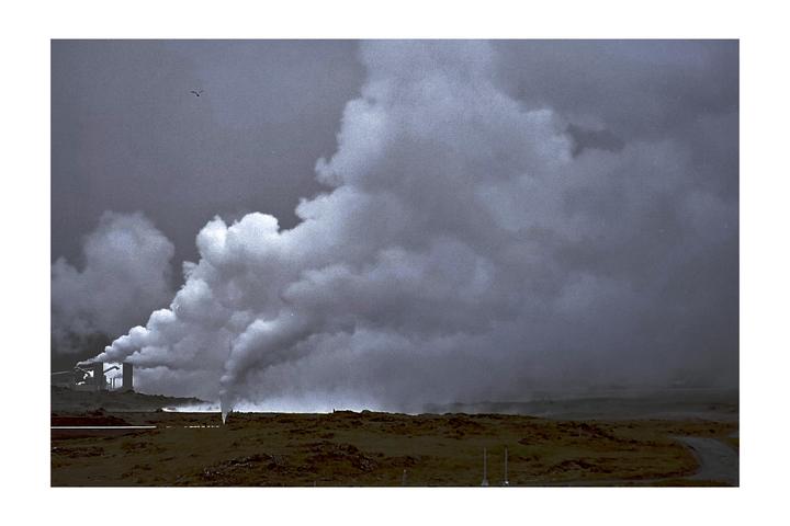 A factory emits large plumes of smoke into a cloudy sky over a barren landscape.
