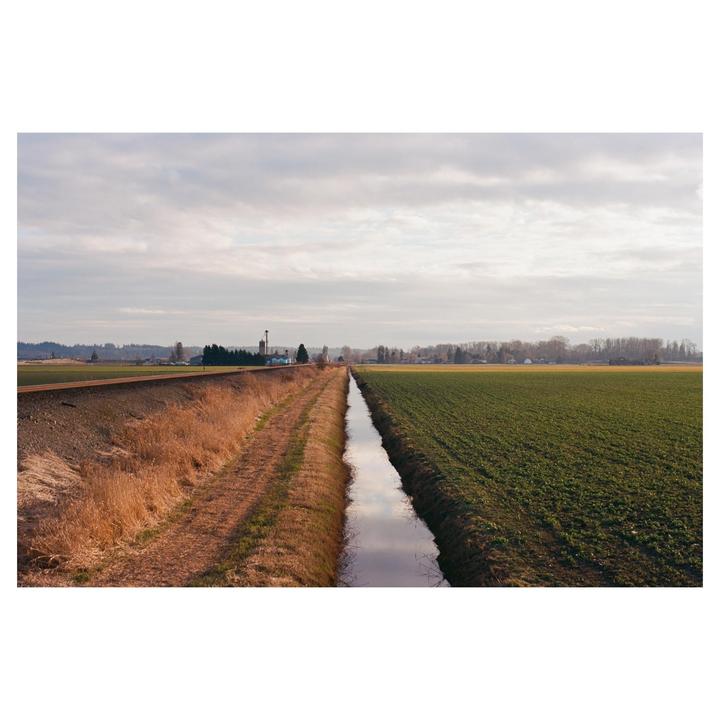 A narrow irrigation ditch separates a field of green crops from a strip of dry grass and a railway line under a cloudy sky.
