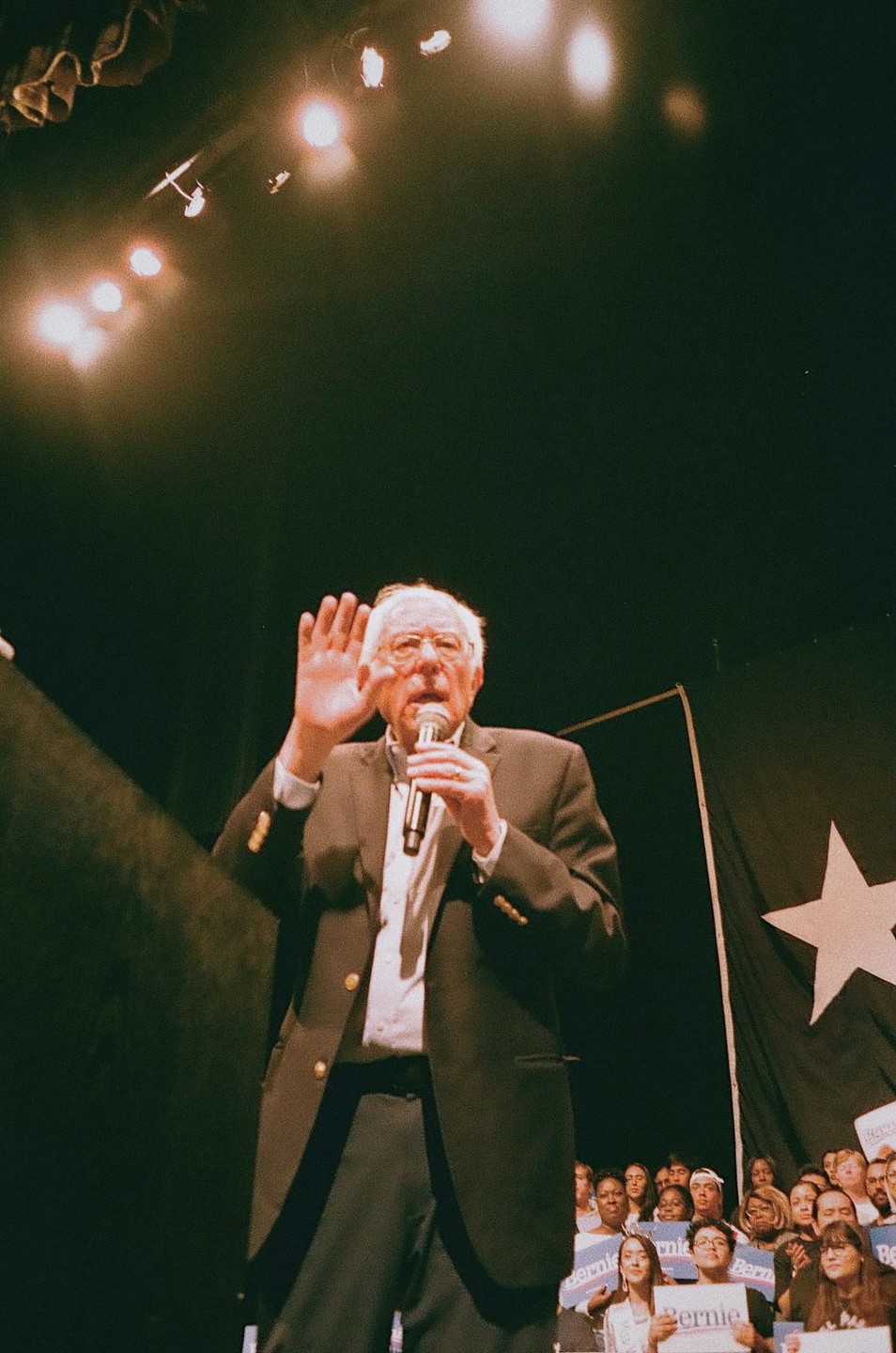 A man in a suit and glasses speaks into a microphone with his hand raised, while a crowd of people watches in the background.