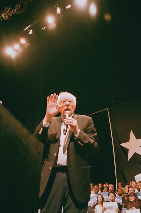 A man in a suit and glasses speaks into a microphone with his hand raised, while a crowd of people watches in the background.