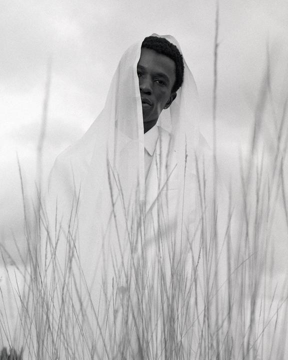 A man with a white veil over his head stands in a field of tall grass.