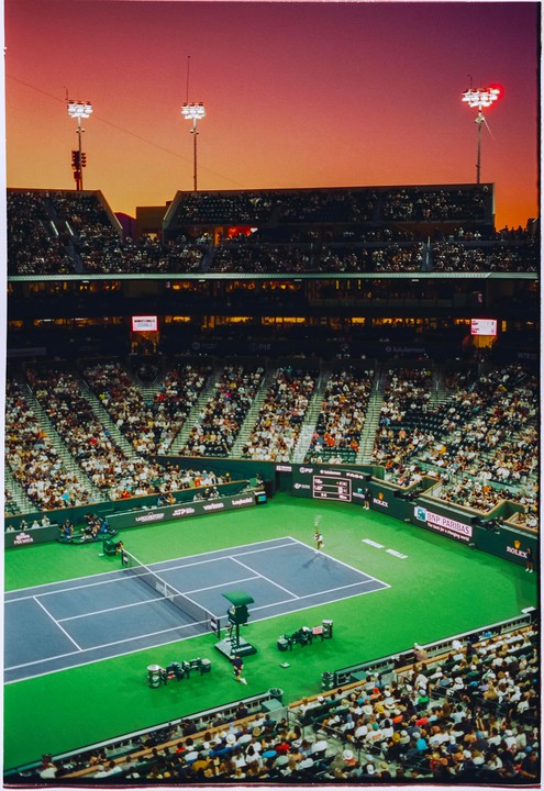 A tennis player serves on a green and blue court in a stadium filled with spectators under a sunset sky.
