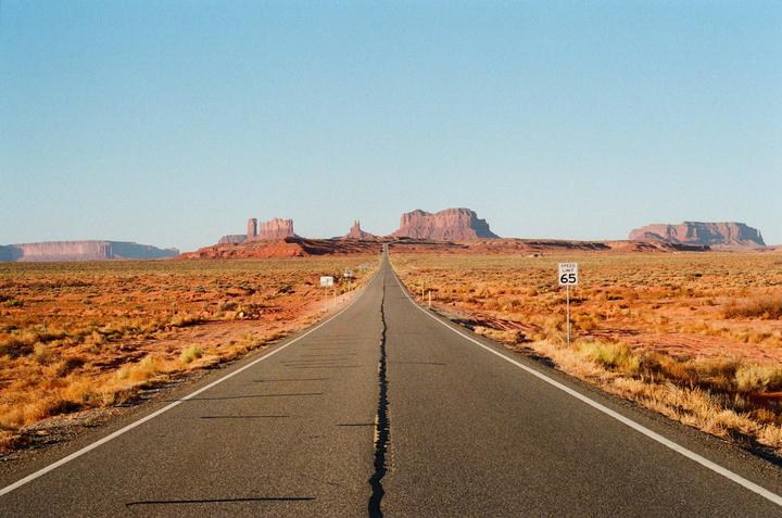 A long, straight road stretches into the distance towards a series of large rock formations under a clear sky.