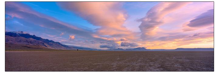 A vast, flat desert landscape stretches towards distant mountains under a sky filled with colorful clouds at sunset.