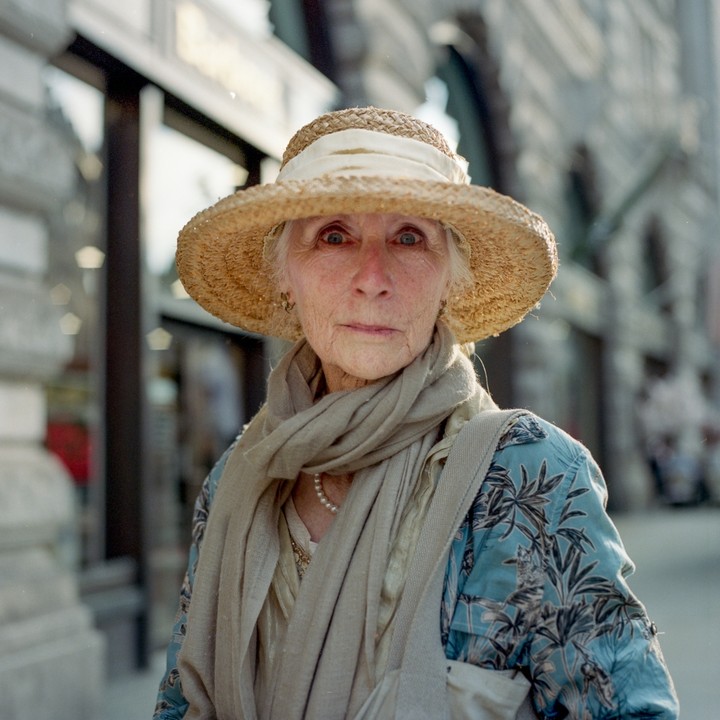 A woman wearing a straw hat and a patterned shirt looks directly at the viewer on a street.