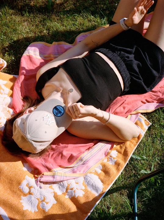 A woman wearing a baseball cap and black clothing is lying on a colorful towel on the grass.