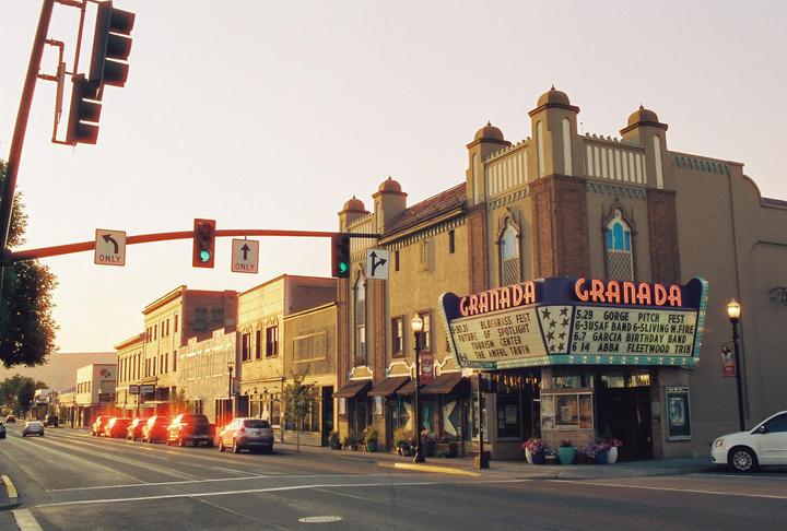 A street with cars and buildings is illuminated by the setting sun, with a prominent theater marquee on the right.