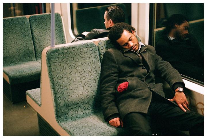 A man sleeps on a train seat next to a window, with another man visible in the background.