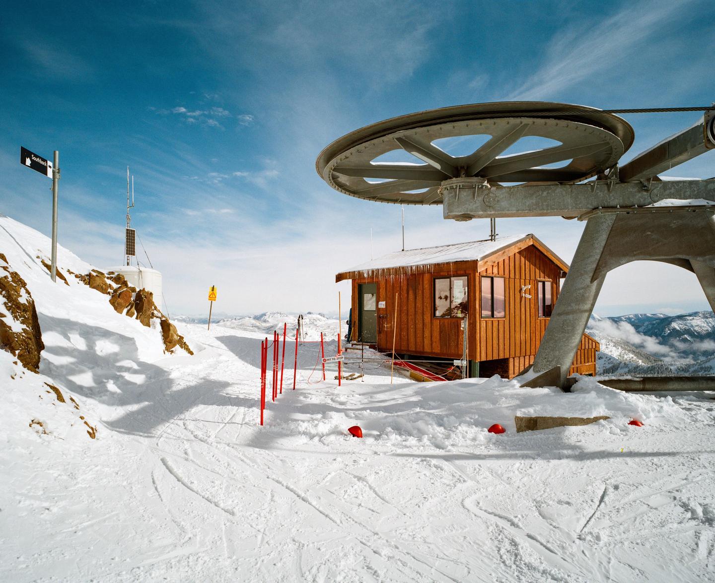 A wooden building with icicles on its roof stands next to a large ski lift mechanism on a snow-covered mountain under a blue sky.