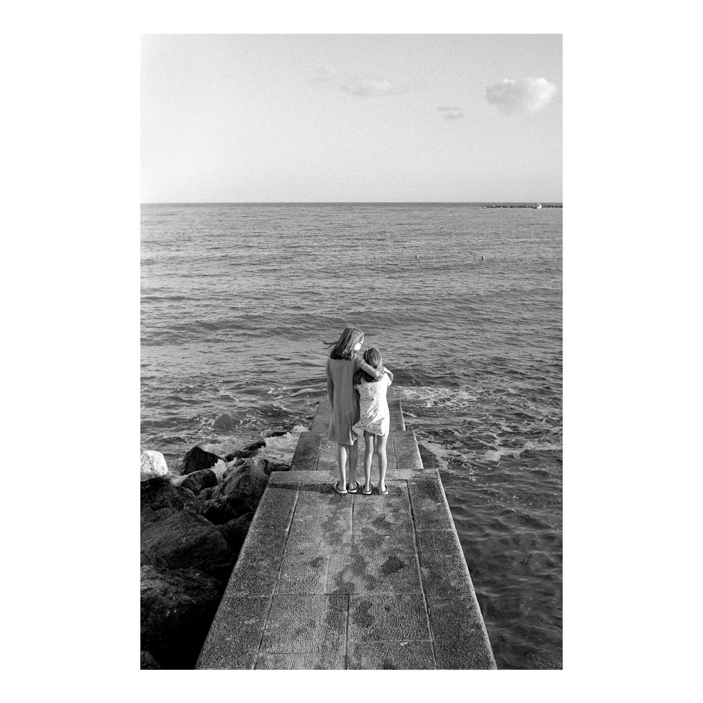 Two girls stand on a concrete pier looking out at the ocean.