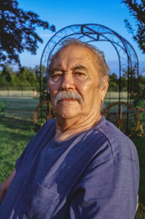 A man with a mustache and a blue shirt is sitting outdoors in front of a decorative metal archway.