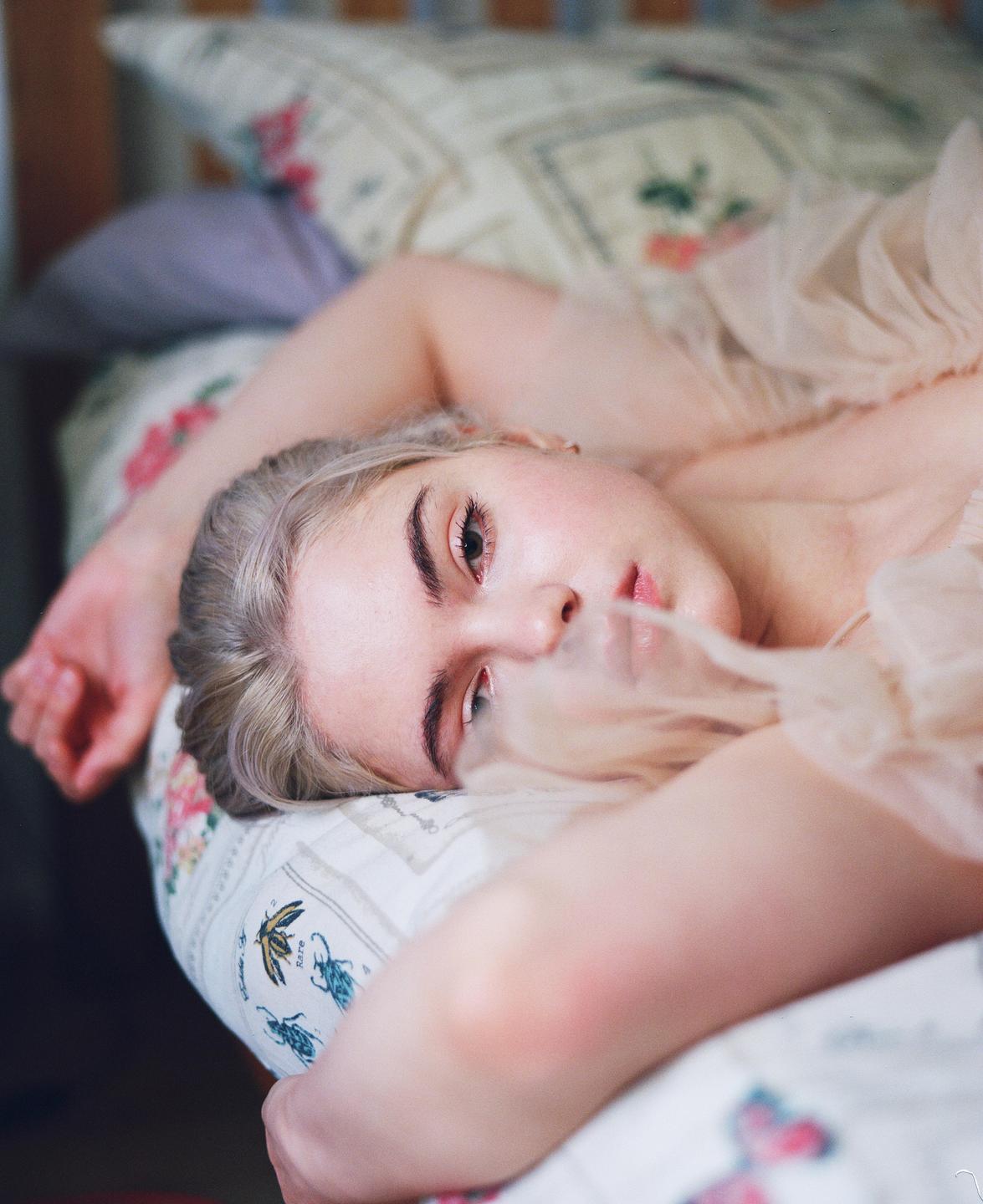 A woman with blonde hair is lying on a bed with patterned bedding, looking directly at the viewer.
