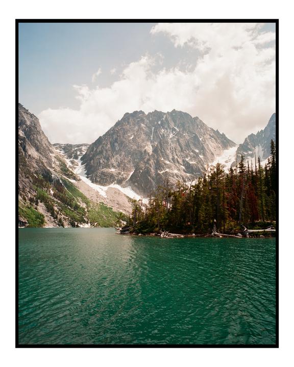 A turquoise lake is surrounded by a forest and snow-capped mountains under a cloudy sky.