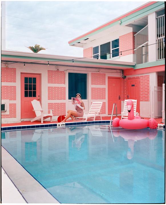 A woman sits by a swimming pool reading a magazine next to a pink flamingo float.