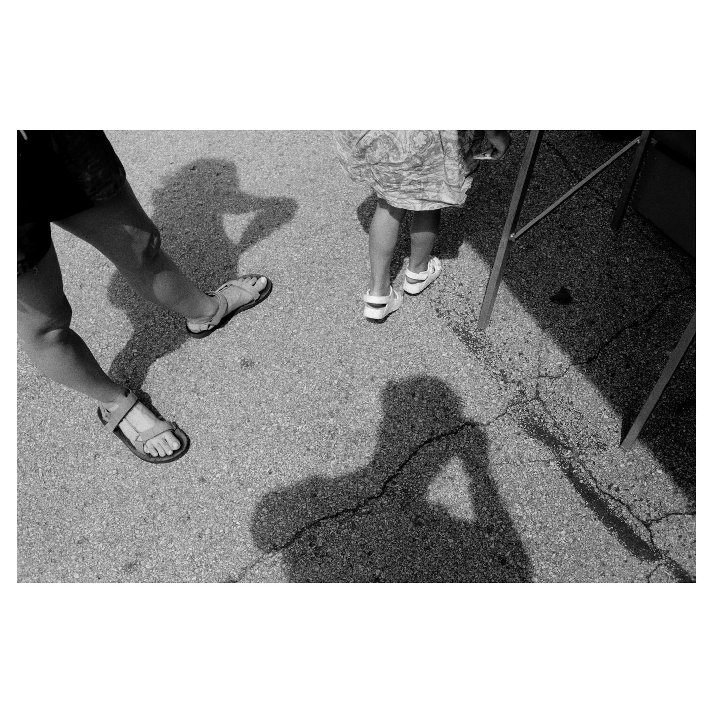 Two people stand on cracked asphalt, casting long shadows, with one shadow appearing to hold a camera.