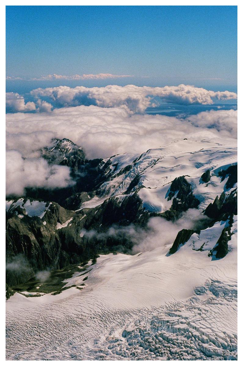 Clouds float above a snow-covered mountain range with a clear blue sky overhead.