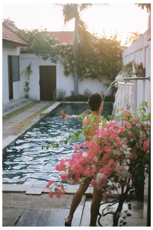 A man showers by a swimming pool with a woman swimming in the background, surrounded by flowering plants.