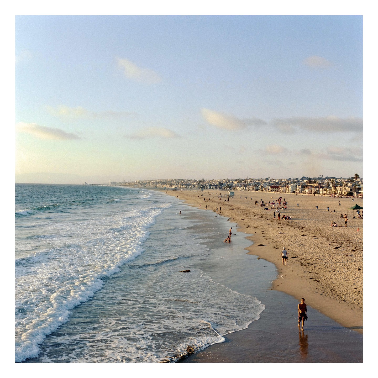 A wide view of Hermosa Beach shows people on the sand and in the water, with a town and pier in the distance.
