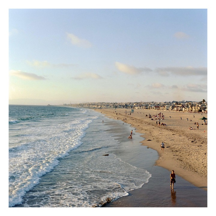 A wide view of Hermosa Beach shows people on the sand and in the water, with a town and pier in the distance.