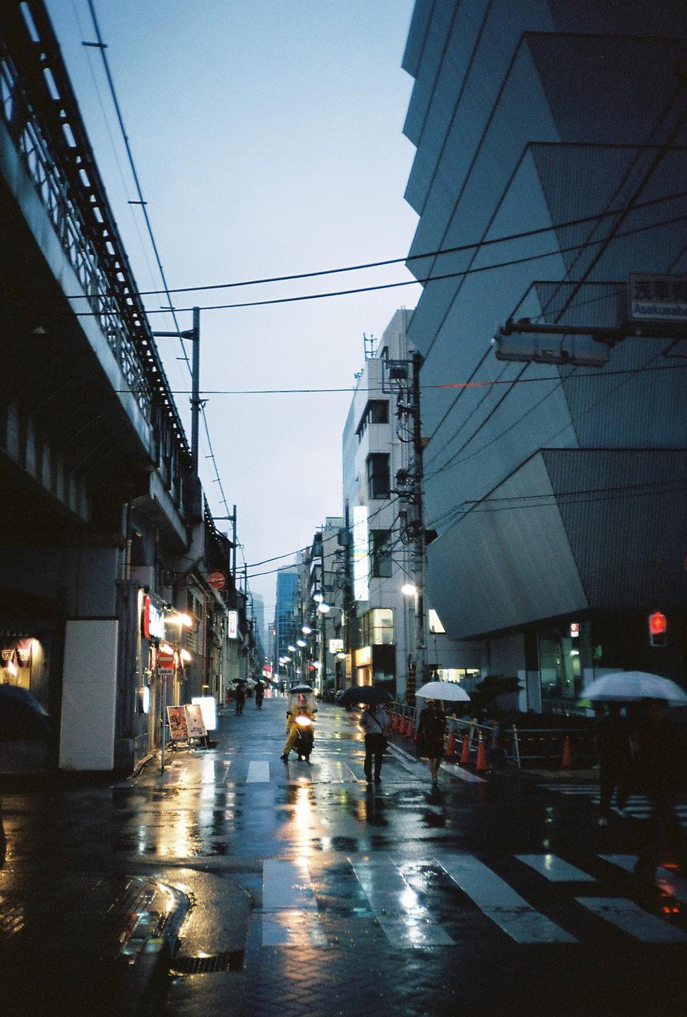 A street in Tokyo is wet from rain, with people walking with umbrellas and lights reflecting on the pavement.