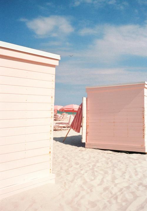 Two pink beach huts stand on a sandy beach with pink umbrellas and lounge chairs in the background under a blue sky.