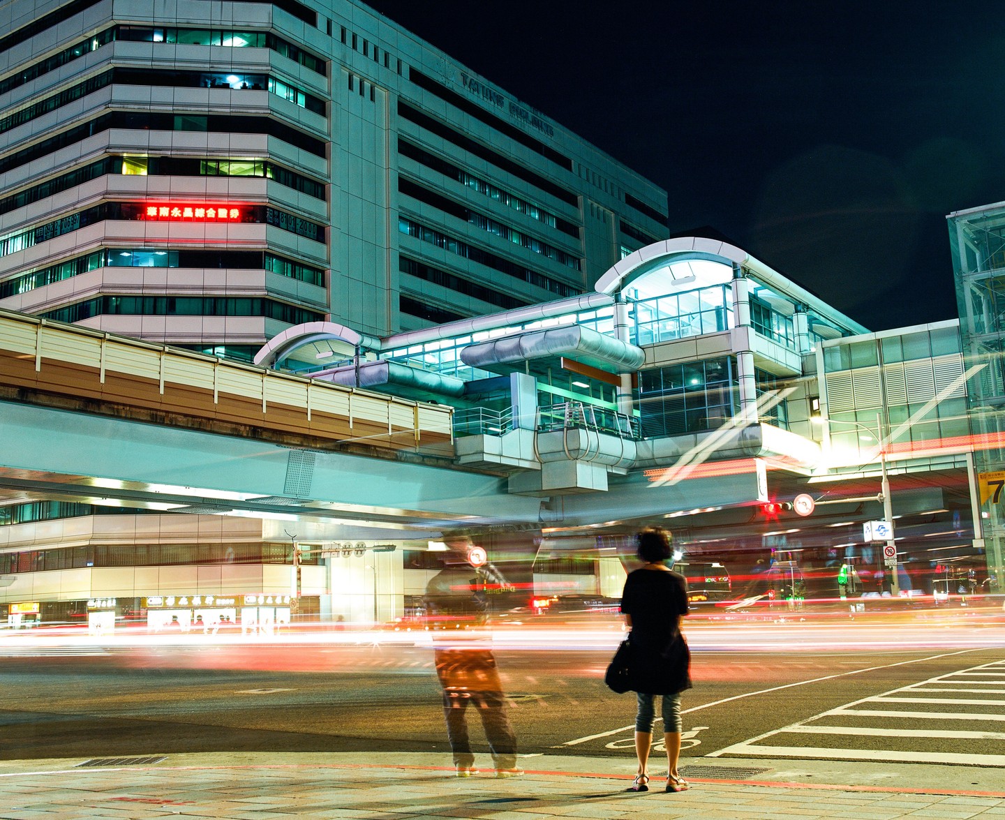 A woman stands on a street corner at night, looking at a building with an elevated walkway and light trails from traffic.