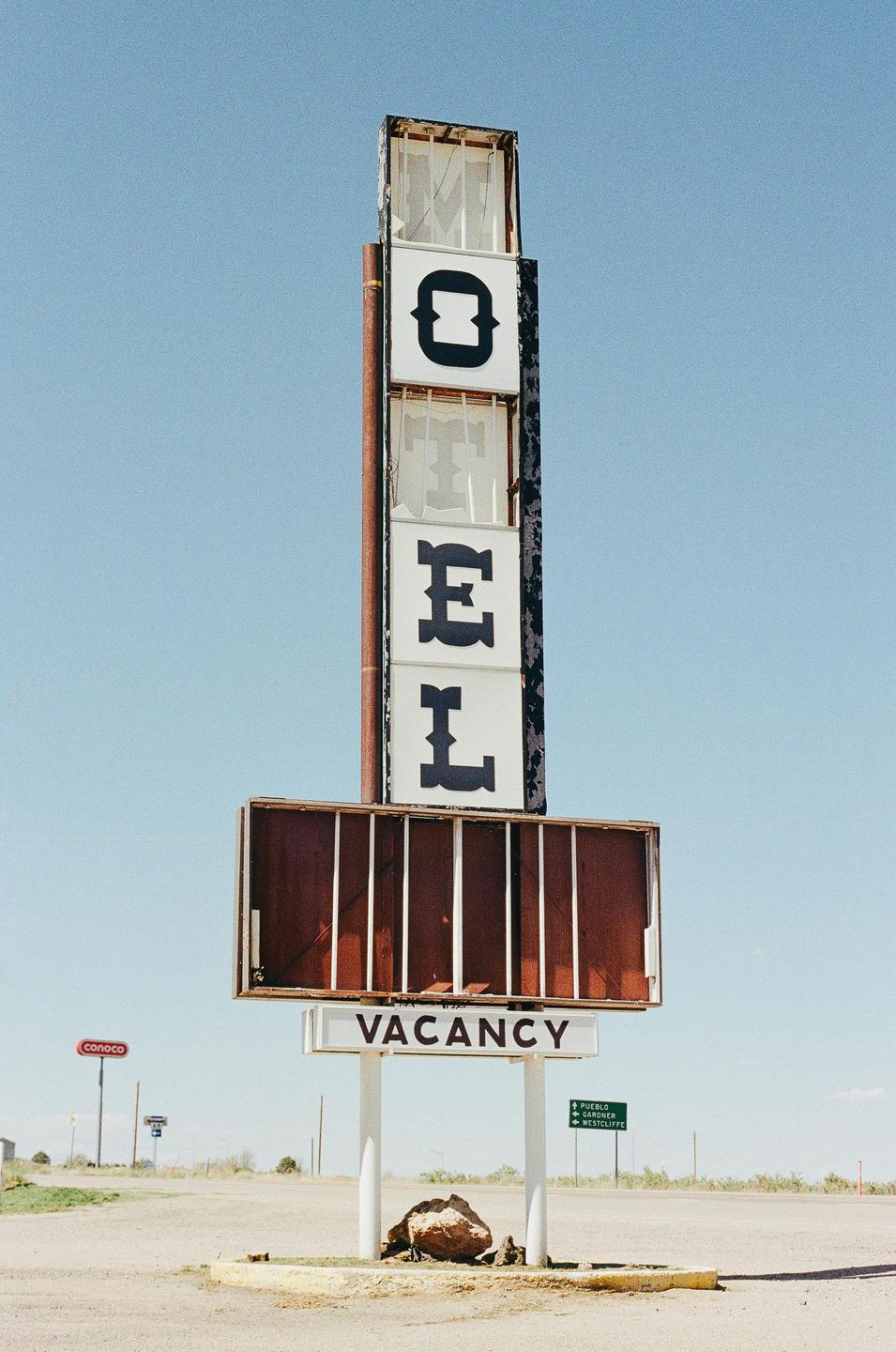 A tall, weathered motel sign with the word "MOTEL" vertically displayed and a "VACANCY" sign below stands against a clear blue sky.