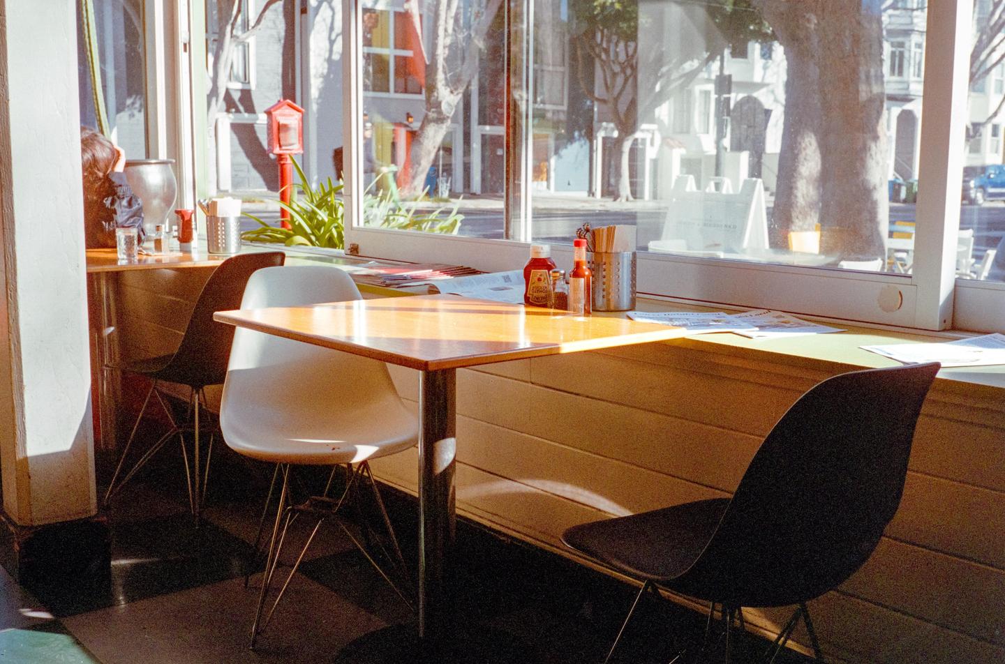 A diner interior with tables and chairs is illuminated by sunlight streaming through large windows.