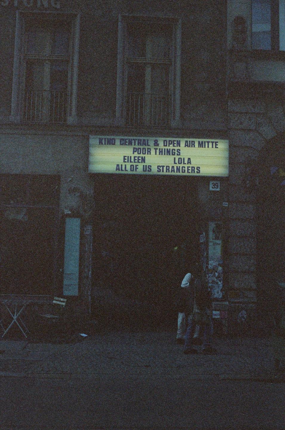 A movie theater marquee advertises several films above a dark entrance where two people stand.