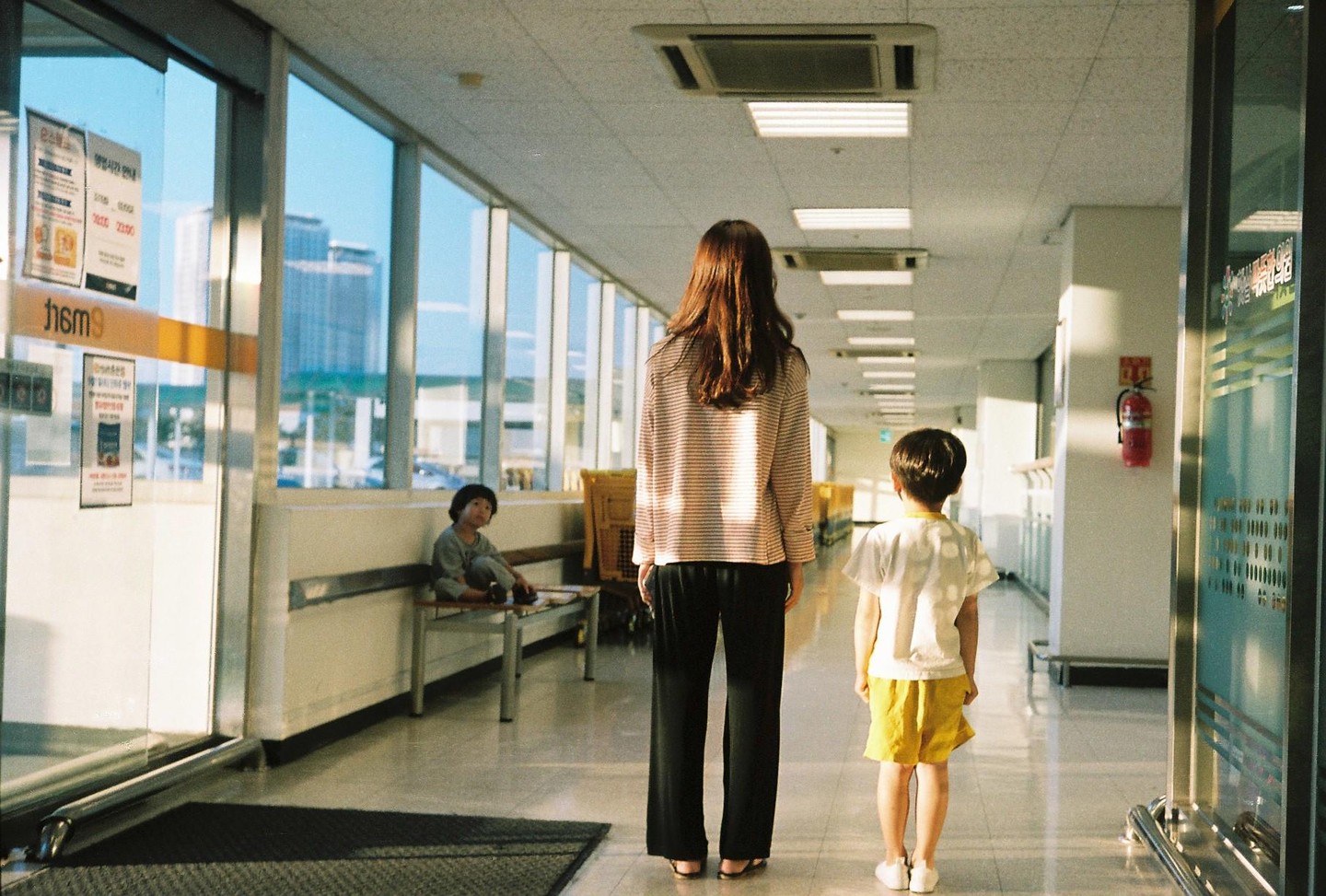 A woman and a child stand in a brightly lit hallway while another child sits on a bench.