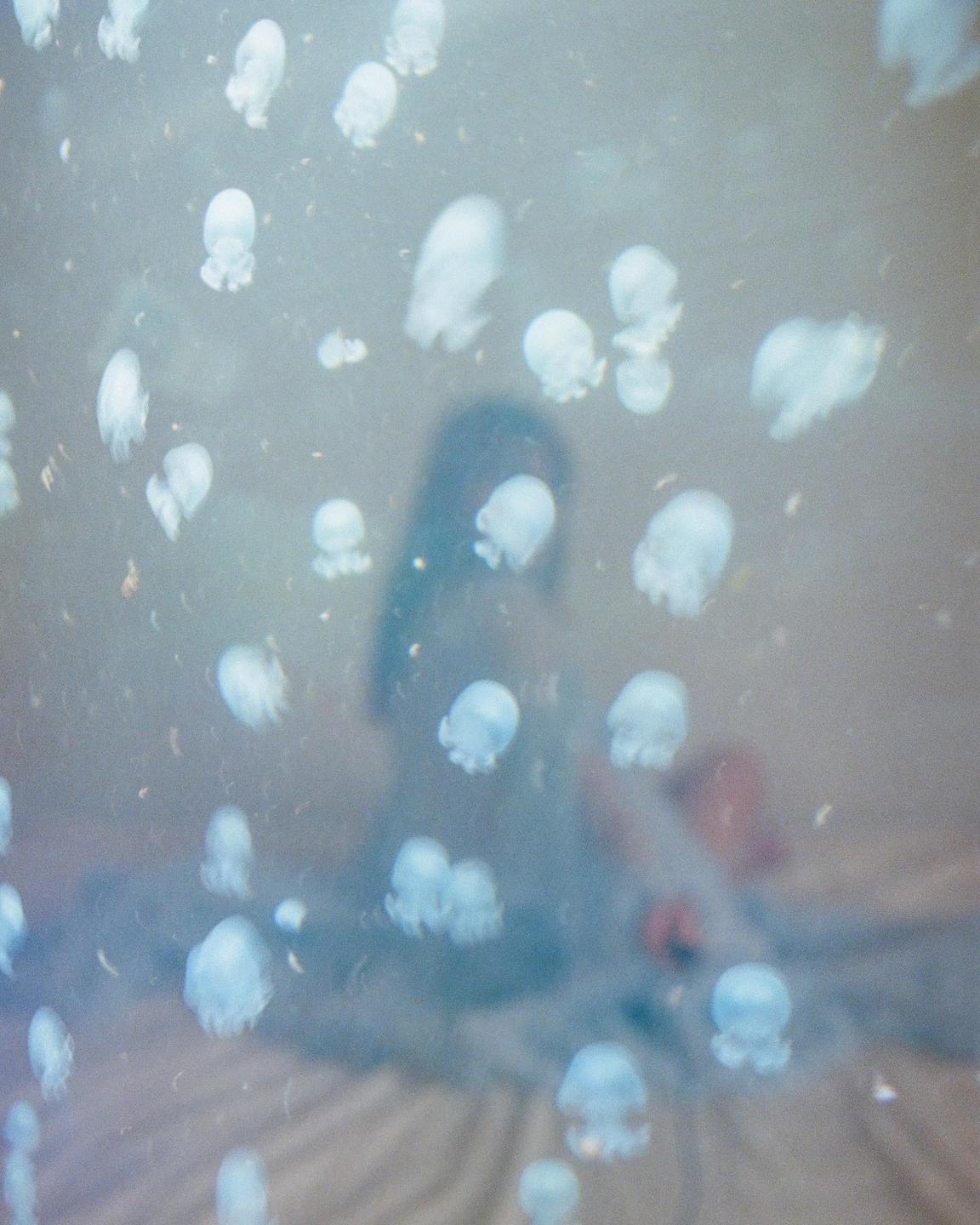 A woman sits on a bed, surrounded by many small, glowing, jellyfish-like forms.