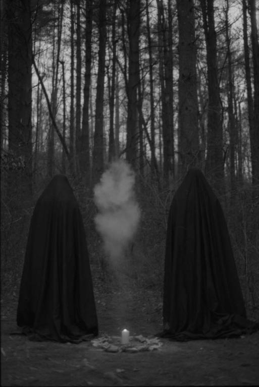 Two figures in dark cloaks stand in a forest with a lit candle and smoke between them.