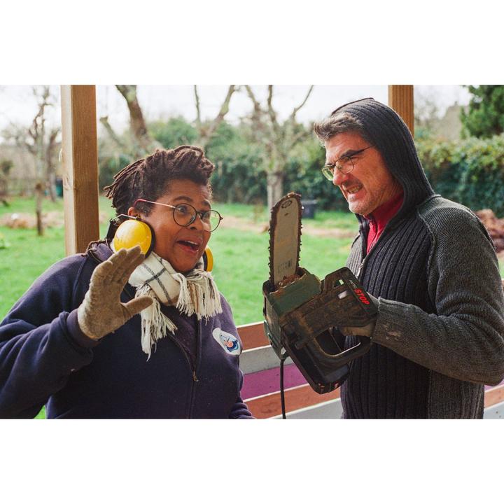 A man holds a chainsaw towards a woman wearing ear protection and a scarf.