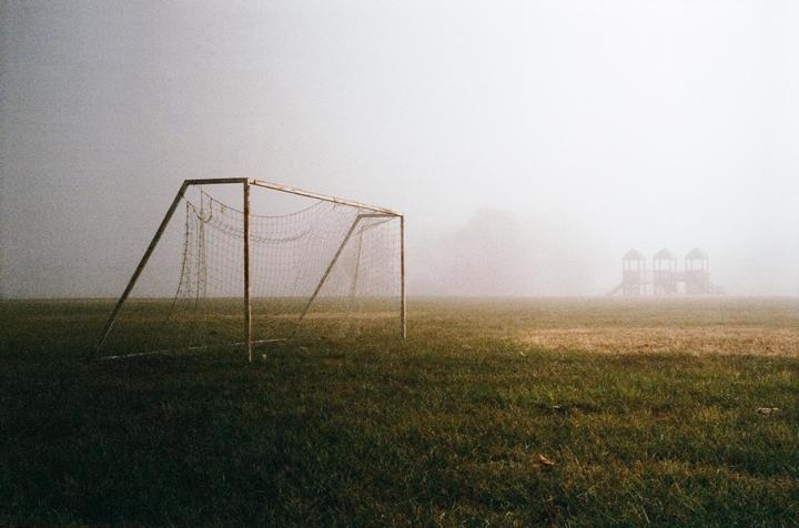 A soccer goal stands on a grassy field with a playground visible in the distance through a thick fog.