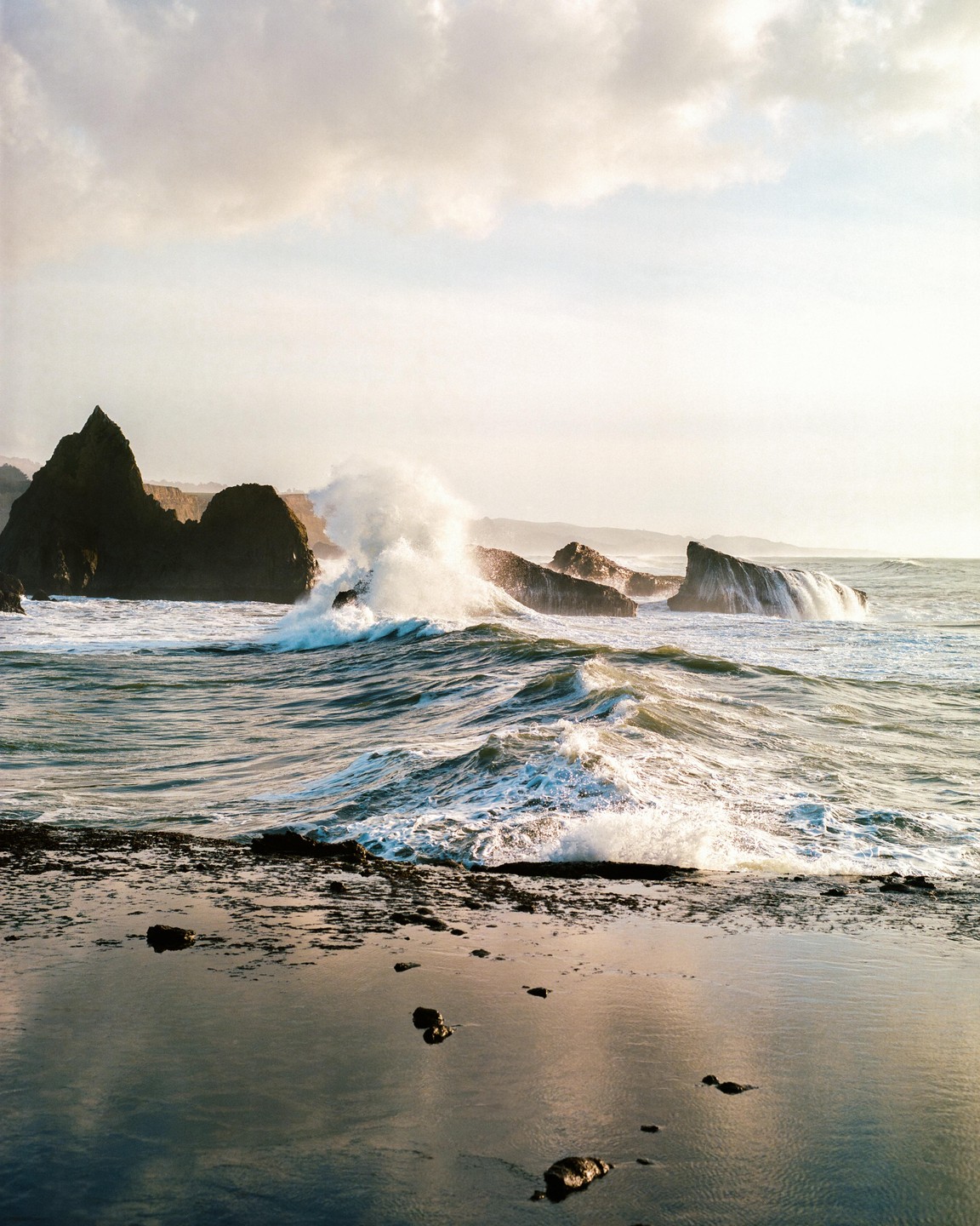 Waves crash against large rocks on a beach under a cloudy sky.