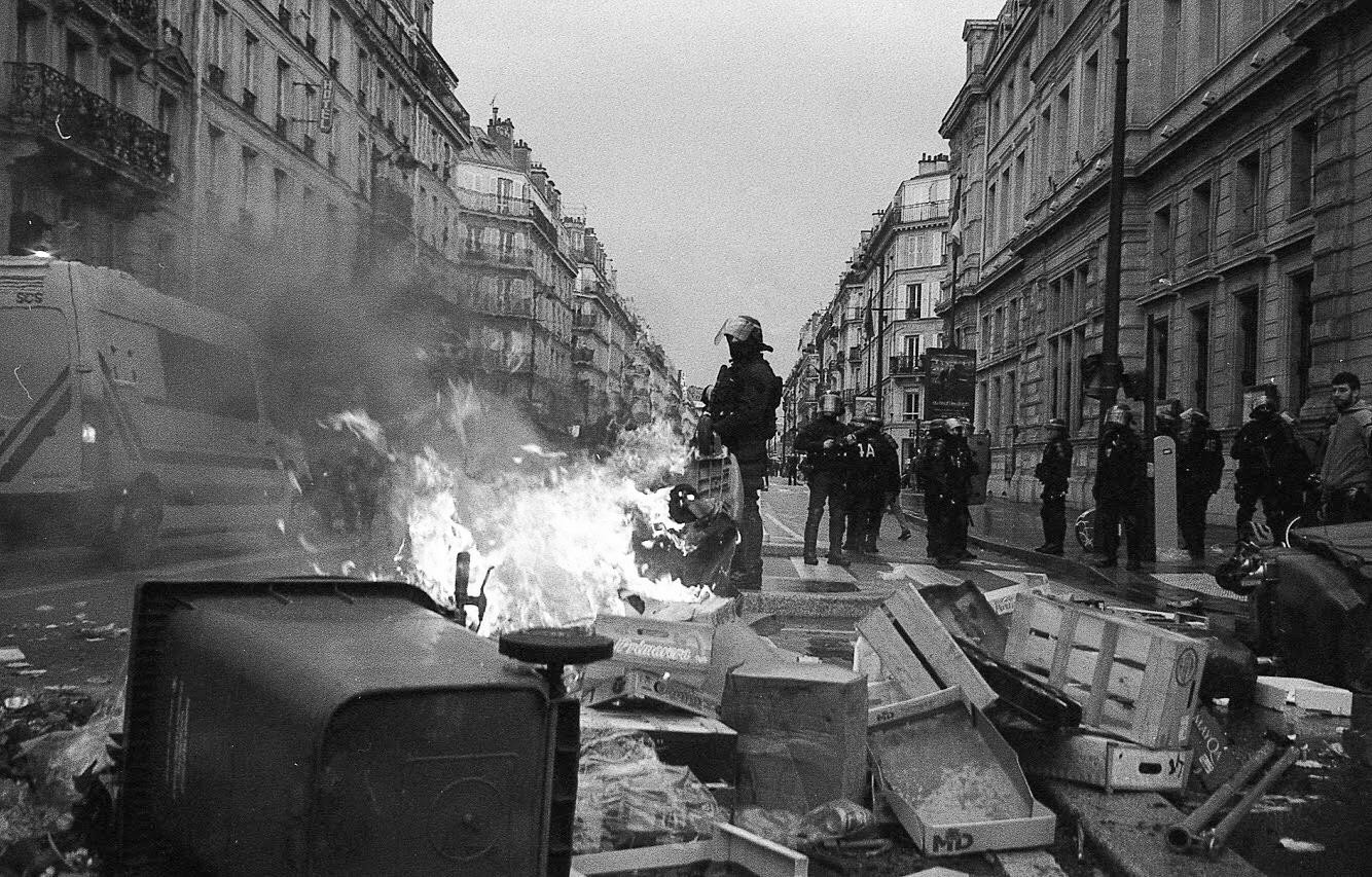 A street scene shows a fire burning in the foreground with police officers standing in the background.