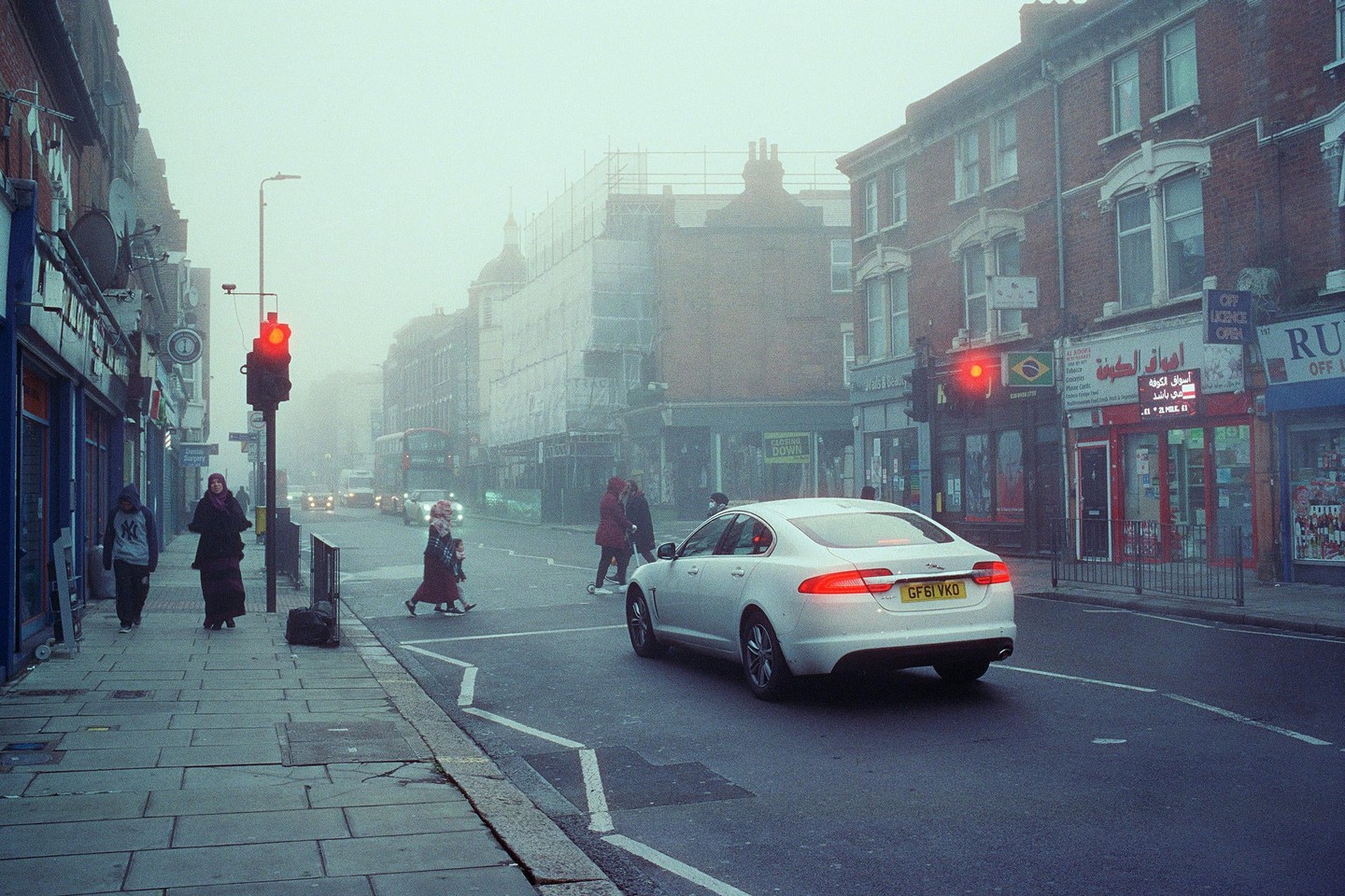 A white car is stopped at a red traffic light on a foggy street with buildings and pedestrians.