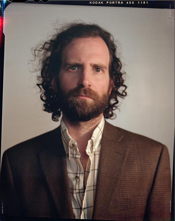 A man with curly hair and a beard wears a brown jacket and a plaid shirt.