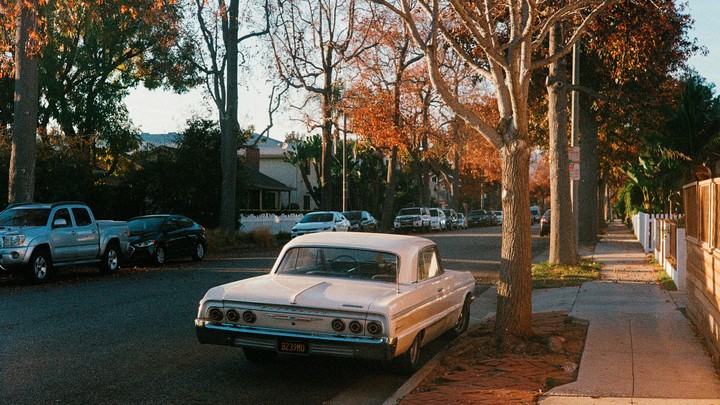 A vintage white car is parked on a residential street lined with trees displaying autumn foliage.