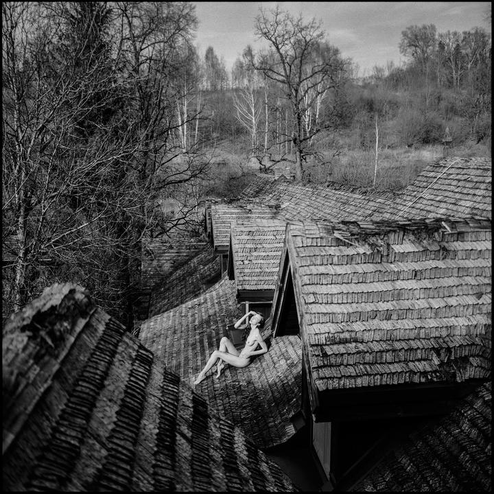 A nude woman is lying on a wooden shingle roof with a forest in the background.