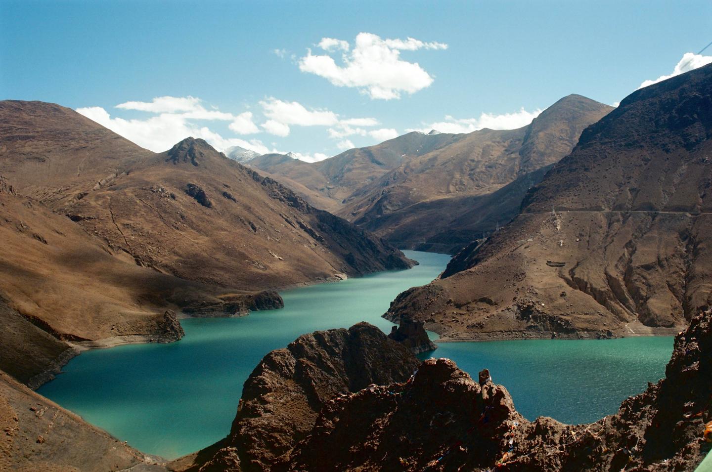 A turquoise river winds through a valley surrounded by arid mountains under a blue sky with scattered clouds.