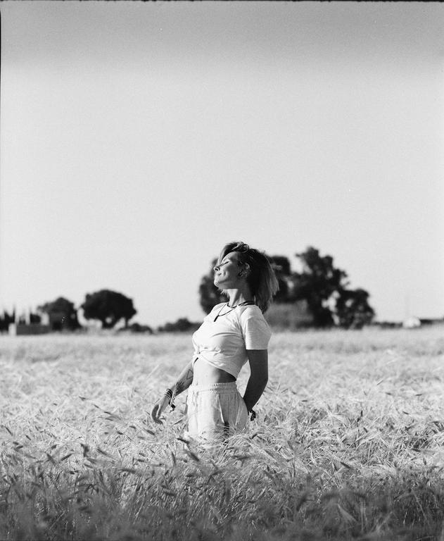 A woman stands in a field of tall grass with her head tilted back and eyes closed.