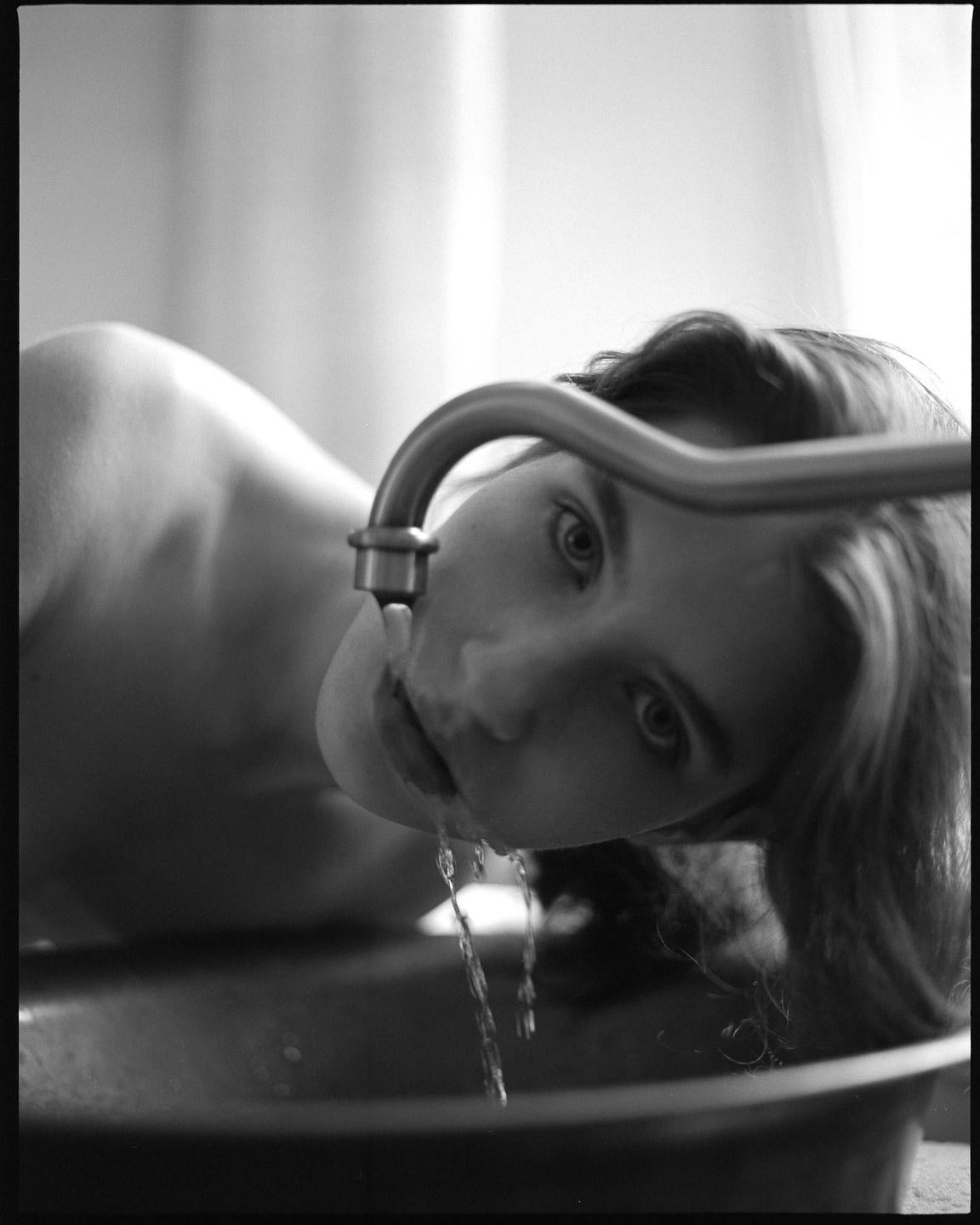 A woman drinks water from a faucet over a basin.